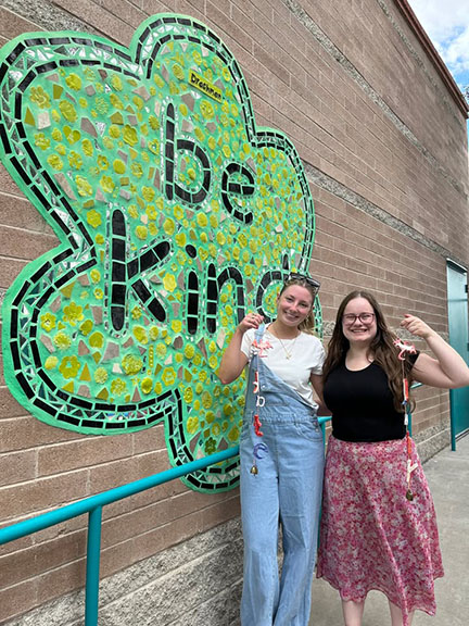 Two young women smile in front of a be kind mural