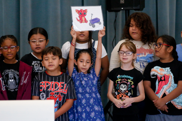 A girl holds up a poster with a baby dragon and a wagon on it, surrounded by her classmates performing