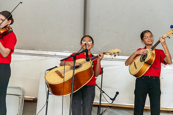 Three girls perform on stage with the mariachi group. Center with a large guitar, left with a violin, right with a traditional guitar