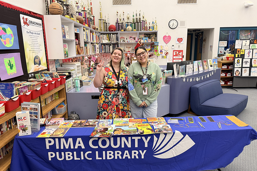 Two women smile, standing behind the Pima County Library table