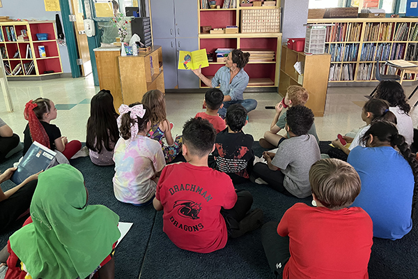 A woman sits on the floor reading to a group of students also sitting on the floor